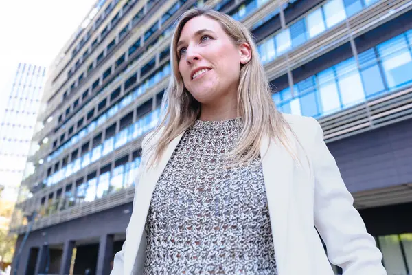 Hafif bir takım elbise giyen, ciddi şekilde kameraya bakan, modern bir ofis binasının önünde duran, liderliği, kararlılığı ve profesyonelliği temsil eden kendine güvenen bir iş kadını.. 