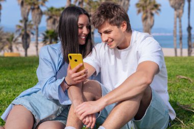 Palmiye ağaçlarının altında deniz kenarında oturan genç çift, birlikte sarı bir akıllı telefona bakarken gülümsüyor, ekrana işaret eden erkek arkadaş, aşkı, seyahat anlarını ve bugün dijital bağlanmayı yakalıyor. 