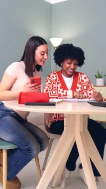 İki genç kadın bir masada birlikte oturup dikey format sahnesinde eğitim, öğrenme, arkadaşlık ve takım çalışmasına odaklanarak kapalı bir ortamda parlak bir şekilde çalışırlar ve gülümserler. 