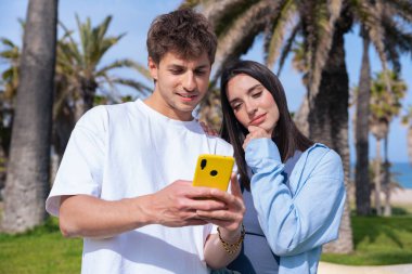 Palmiye ağaçlarının altında deniz kenarında oturan genç çift, birlikte sarı bir akıllı telefona bakarken gülümsüyor, ekrana işaret eden erkek arkadaş, aşkı, seyahat anlarını ve bugün dijital bağlanmayı yakalıyor. 