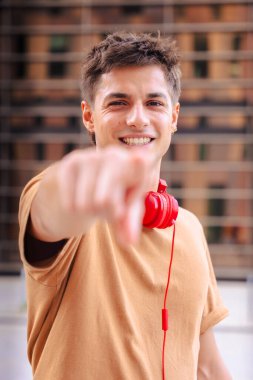 Kendine güvenen, gülümseyen ve doğrudan kameraya bakan, boynuna kırmızı kulaklık takmış, kahverengi tişört giymiş, dışarıda dikey duran genç bir adam.