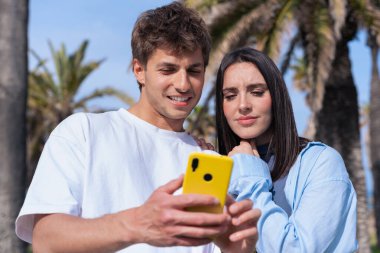 Palmiye ağaçlarının altında deniz kenarında oturan genç çift, birlikte sarı bir akıllı telefona bakarken gülümsüyor, ekrana işaret eden erkek arkadaş, aşkı, seyahat anlarını ve bugün dijital bağlanmayı yakalıyor. 
