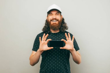 Kapalı portre genç neşeli sakallı adam beyaz şapka giyen ve gri arka plan üzerinde akıllı telefon ekranı gösteren.