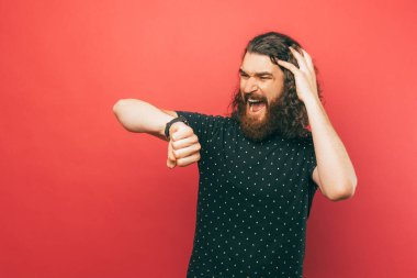 Şok olmuş genç sakallı bir adamın pembe arka plan üzerinden saate bakarken çekilmiş fotoğrafı..