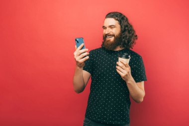 Uzun saçlı, mutlu ve sakallı bir adamın fotoğrafı. Akıllı telefon kullanıyor ve pembe arka planı incelemek için elinde bir fincan kahve tutuyor..