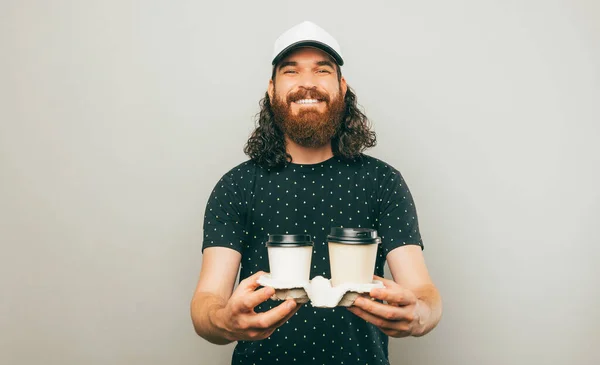 stock image Photo of cheerful bearded hipster man wearing white cap and holding two cups of coffee to go over grey background.