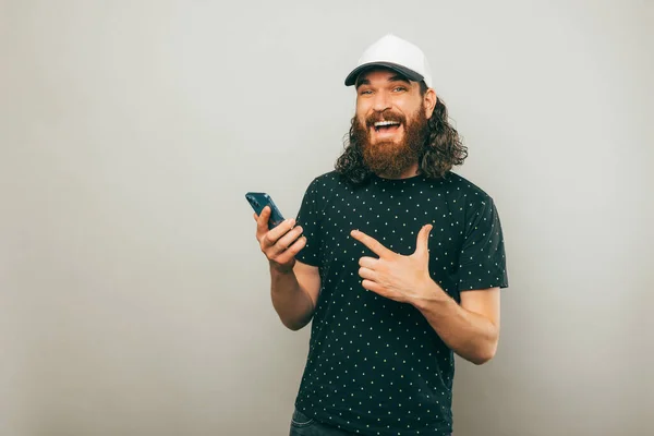 Portrait Joyful Bearded Man Wearing White Cap Pointing Smartphone Gray — Stock Photo, Image