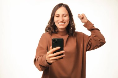 Gülümseyen, akıllı telefona bakan ve zaferi kutlayan neşeli genç bir kadının fotoğrafı..