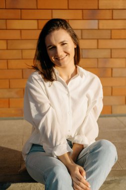 Duvarın yanındaki parlak güneşte kameraya bakıp gülümseyen genç bir kadının dikey portresi..