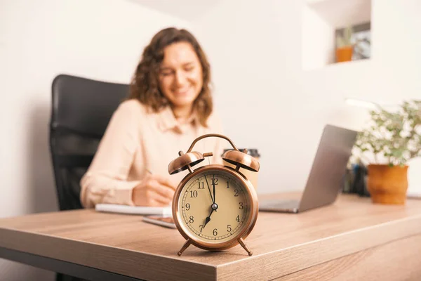 Close up photo of alarm clock on office table and woman making notes in agenda.