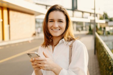 Açık havada genç ve güzel bir kadının portresi kameraya bakıyor ve elinde akıllı telefon, şehir arka planı.