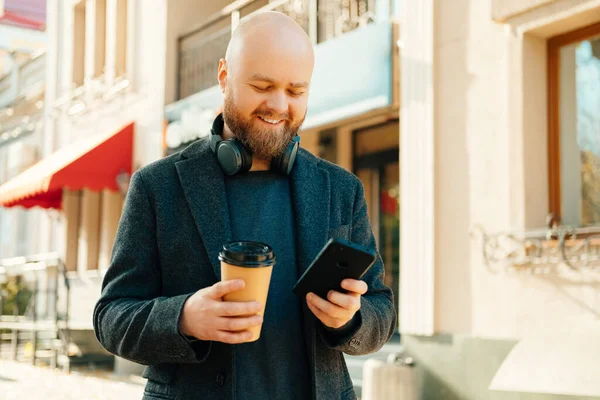 Cheerful bald bearded man is browsing in the phone while drinking to go coffee on the street.
