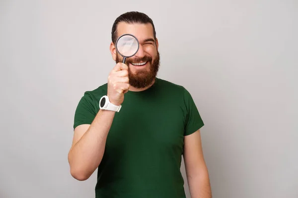 Portrait of a bearded man saying I am looking for you through magnifying glass in a grey studio.