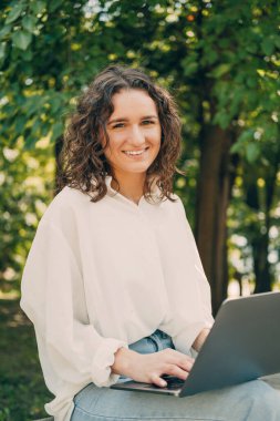 Bilgisayarda çalışan genç bir kadının fotoğrafı. Parkın dışında ve gülümsüyor..
