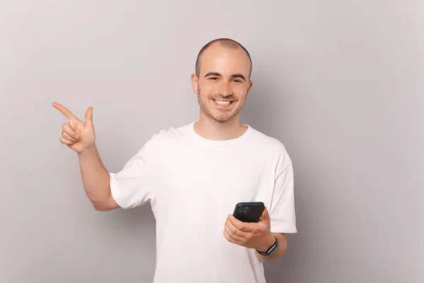 Радісний Молодий Чоловік Вказує Осторонь Сірий Простір Копіювання Тримаючи Телефон — стокове фото