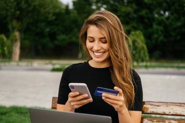 Geniş gülümseyen kadın dışarıda bankta otururken telefonunu ve kredi kartını kullanıyor..