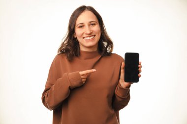 Gülümseyen sevimli genç bayan elinde tuttuğu telefon ekranını işaret ediyor. Stüdyo çekimi beyaz arkaplan üzerinde.