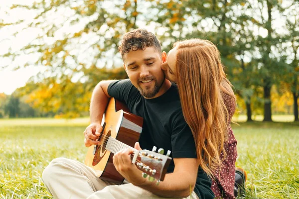 若いです女性接吻彼女の男彼再生ザギター座っている間に草の上に公園 — ストック写真