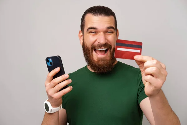 Wide smiling man loves mobile banking experience with his new credit card. Studio shot over grey background.