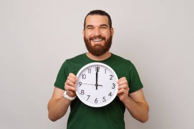 Geniş gülümseyen sakallı adam açık gri arka planda zamanı temsil eden yuvarlak beyaz bir saat tutuyor..