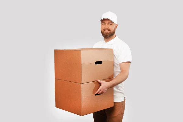 Image of young delivery man in white uniform holding two big boxes and looking at camera over grey background.