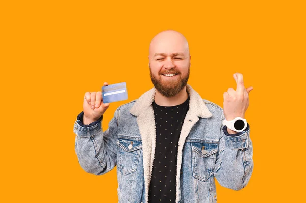 Excited young bearded bald man crossing fingers for a wish while holding card over yellow background.