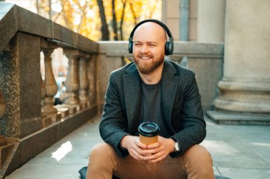 Young bald man listens to a podcast while enjoying a cup of coffee outdoors sitting on some stairs.
