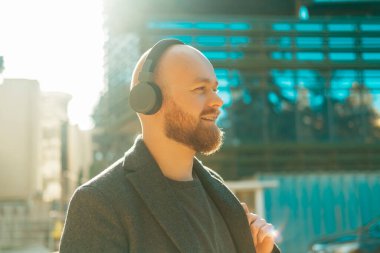 Handsome bald bearded man walks in the sunlight while wearing headphones.