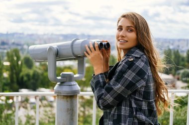 Gorgeous woman holds the sightseeing binoculars in order to look through them at the beautiful city ahead.