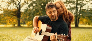 In love couple are sitting on the grass while man is playing the guitar in a green park.