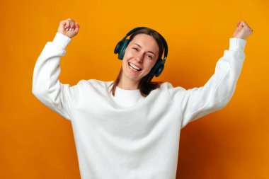 Young woman has fun while dancing and listening to the music in headphones over yellow background.
