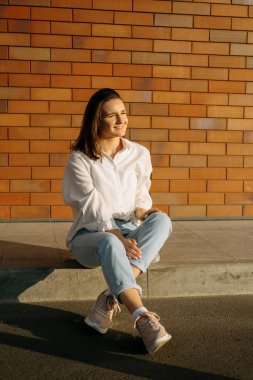 Young smiling woman is sitting in the sunlight outdoors near a brick wall. Vertical shot.