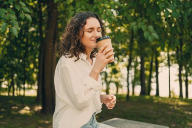 Young curly woman enjoys to go coffee with eyes closed in a green park while taking a break from work.