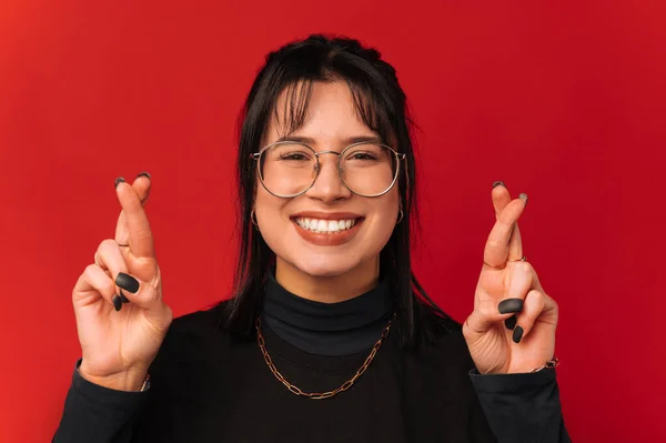 Cheerful woman with black hair is crossing fingers while wishing for something over red background.