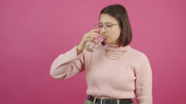 Footage of young woman drinking fresh water from glass over pink background.