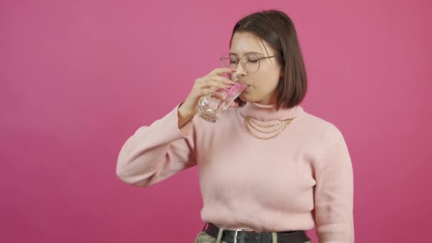 一位年轻女子在粉红的背景上用玻璃杯喝水的镜头 — 图库视频影像