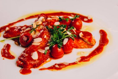 White plate with some fish fillet in cherry tomato sauce decorated with greens from a restaurant.