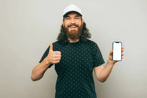 Handsome smiling curly man is showing screen of the phone and thumb up over light grey background.