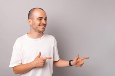 Handsome young man is pointing aside with two hands over grey background in a studio.