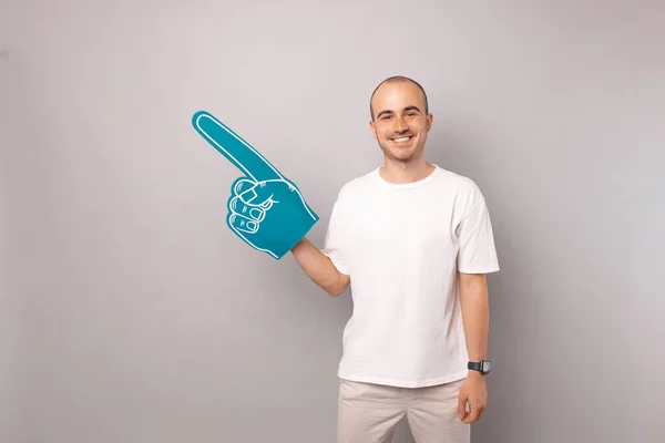 Handsome balding man is wearing foam fan cheering finger glove over grey background.