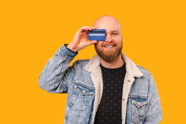 Smiling bearded balding man covers playful one eye with a blue credit card over yellow background.