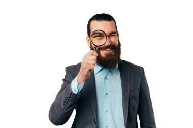Handsome smiling man wearing jacket is looking through magnifying glass over white background.