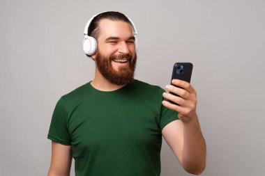 Handsome smiling bearded man wearing headphones is looking at the phone over grey background.