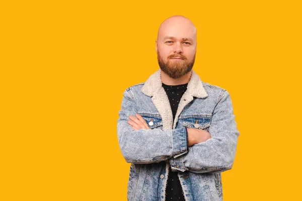 Handsome bearded bald man wearing jeans jacket is staying with arms crossed over yellow backdrop.