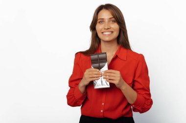Cheerful young woman wearing red shirt holding an opened chocolate bar over white background.