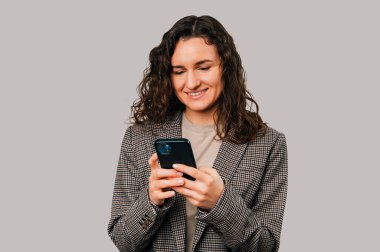 Smiling curly woman is typing a message or surfing the internet over the phone over grey background.