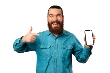 Young cheerful bearded man pointing at his phone screen with copy space over white background.