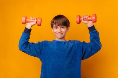 Strong handsome boy is lifting up two orange dumbbells over yellow backdrop in a studio.