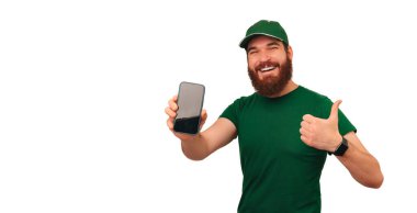 Cheerful delivery man in green shows the screen of his phone and a thumb up over white background.