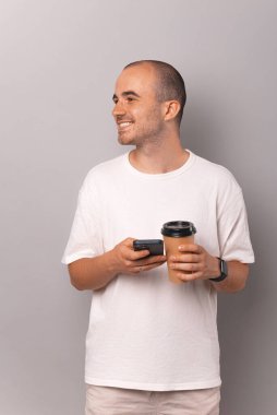 Vertical shot of a handsome young bald man holding phone and to go coffee cup over grey background.
